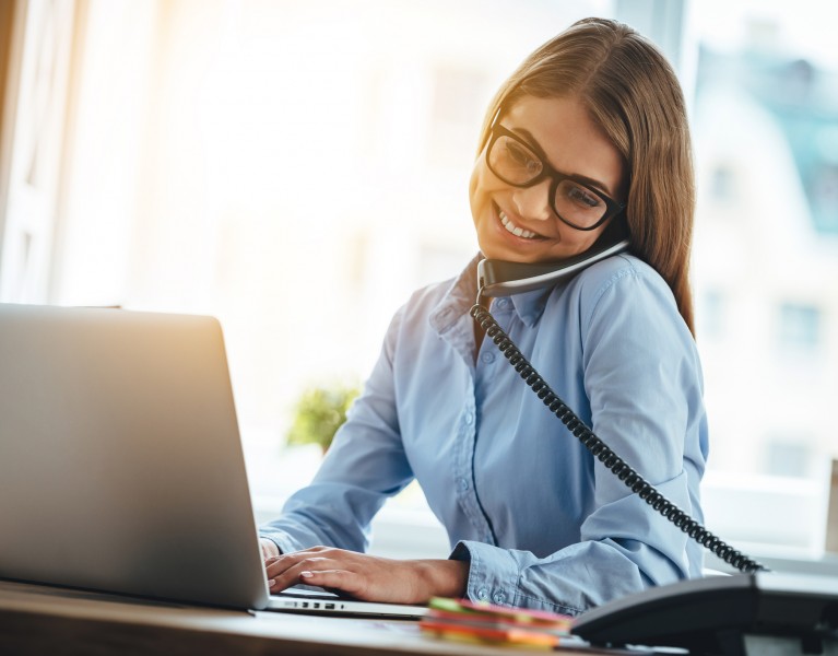 Cheerful young beautiful woman in glasses talking on the phone and using laptop with smile while sitting at her working place